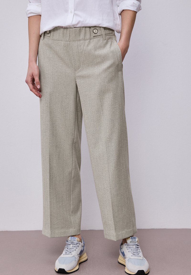 Osoba nosící béžové široké kalhoty s rybí kostí, bílou košili s vyhrnutými rukávy a šedomodré tenisky, stojící s jednou rukou v kapsě.