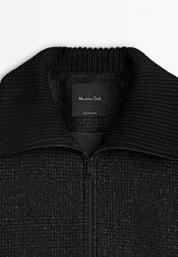 Svart zip-up tröja med en tjock ribbad krage, texturerad tyg, och en etikett inuti som visar "Massimo Dutti" och "Tillverkad i Rumänien."