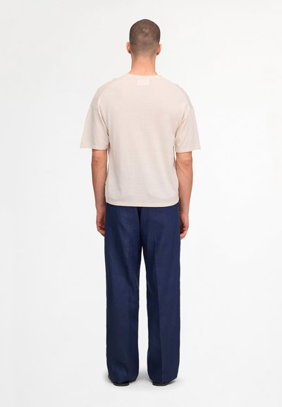 Homme debout de dos, portant une chemise beige clair unie à manches courtes et un pantalon ample bleu marine foncé sur fond blanc.