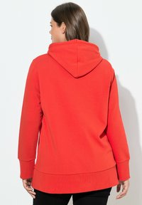 Sudadera roja hecha de una tela suave, con una gran capucha, mangas largas y un dobladillo trasero ligeramente alargado, diseñada para la comodidad.