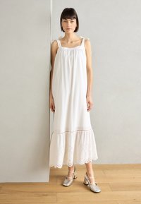 Vestido de lino blanco con un dobladillo bordado en forma de campana y tirantes en los hombros. Combinado con zapatos de tacón bloque metálico plateado. Fondo de suelo de madera natural.