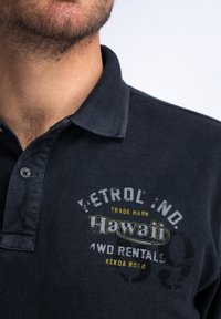 Granatowa koszulka polo wykonana z teksturowanej tkaniny. Posiada kołnierzyk, zapięcie na dwa guziki oraz nadruk "Hawaii" z różnymi akcentami i logo.