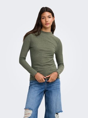 Kvinde med langt mørkt hår iført en tætsiddende olivengrøn ribstrikket langærmet bluse og revnede blå jeans mod en ensfarvet lys baggrund.