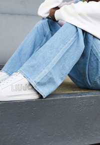 Valkoiset tennarit, joissa on "LIUJO"-logo, yhdistettynä vaaleansinihin leveälahkeisiin farkkuihin. Jalkineiden sileä pinta tuo kontrastia farkkukankaan kanssa.