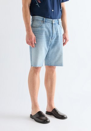 Άνδρας που φοράει ανοιχτόχρωμα τζιν σορτς με ακατέργαστο τελείωμα, μπλε κοντομάνικο πουκάμισο και μαύρα δερμάτινα παπούτσια χωρίς κορδόνια, στέκεται σε λευκό φόντο.