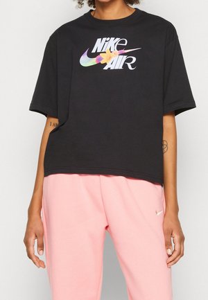 Osoba nosząca czarny krótki t-shirt Nike Air z kwiatowym wzorem na logo oraz jasnoróżowe spodnie dresowe Nike.