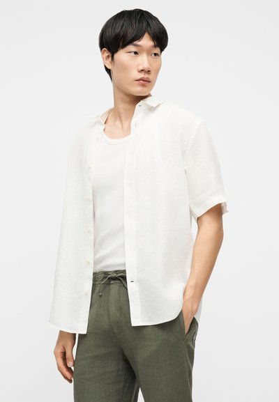 Νέος άνδρας που φορά λευκό ανοιχτό κοντομάνικο πουκάμισο πάνω από λευκό φανελάκι και χακί παντελόνι με κορδόνι, κοιτάζει προς τα πλάγια.