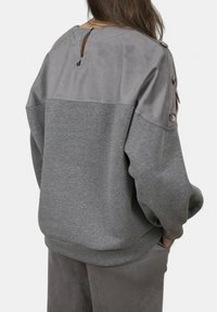 Donna che indossa un maglione grigio largo con parte superiore in suede sulla schiena e dettagli con bottoni sulle maniche, in piedi con le mani nelle tasche dei pantaloni, rivolta di spalle.