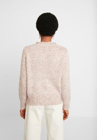 Vaaleanpunainen neulottu villapaita, jossa on teksturoitu kangas, pyöreä pääntie ja resorit hihansuuissa. Päällä kermaiset mantereiset housut, nähtynä takaa.