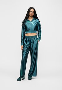 Γυναίκα που φοράει γυαλιστερή τυρκουάζ φόρμα Puma με κοντό μπουφάν με φερμουάρ, παντελόνι με φαρδιά μπατζάκια και ταιριαστά αθλητικά παπούτσια, στέκεται μπροστά σε απλό λευκό φόντο.