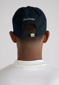 Navy blue baseball cap with "Prohibited" embroidered in white on the back. Adjustable strap at the rear. Worn with a white shirt.