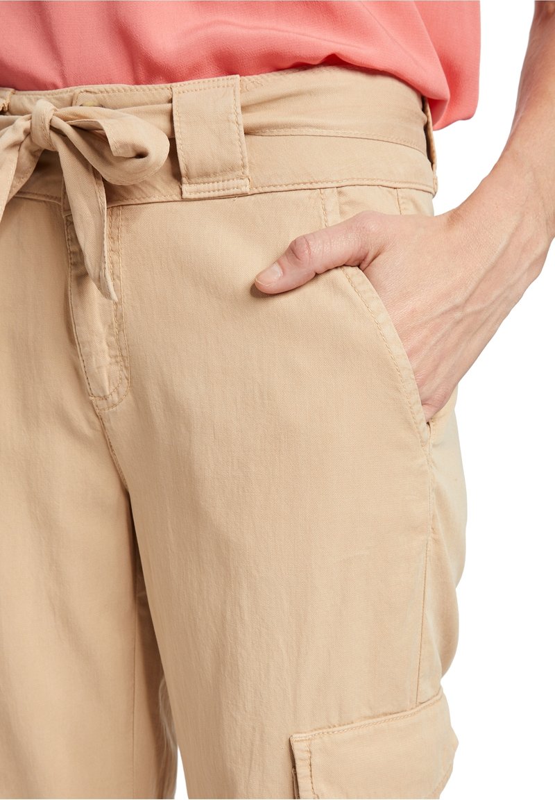 Personne portant un pantalon beige avec une ceinture nouée, la main dans la poche avant, et un haut rose corail visible au niveau de la taille.