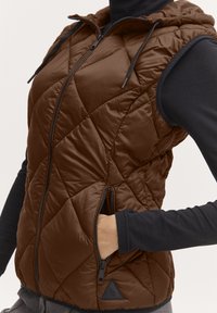 Gilet matelassé marron avec capuche, fermeture éclair à l'avant et poches latérales. Présente une finition texturée et un détail de logo triangulaire sur l'ourlet.