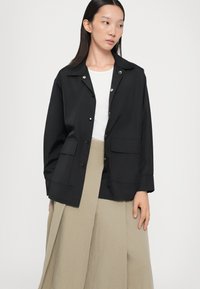 Giacca nera leggera con colletto classico, bottoni a scatto e due tasche frontali, indossata sopra un top bianco e una gonna beige.