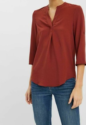 Vrouw draagt een roestkleurige blouse met lange mouwen en V-hals, gecombineerd met een blauwe spijkerbroek, staand tegen een effen lichte achtergrond.