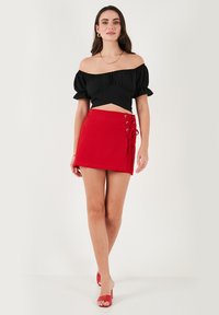 Zwarte off-the-shoulder top met pofmouwen, gedragen met een rode minirok met vetersluiting en metalen oogjes aan de zijkant.
