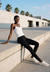 Woman in white tank top, black leggings, and white sneakers sitting on concrete ledge outdoors with palm trees and modern architecture behind.
