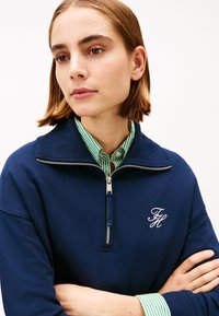 Μπλε ναυτικό φούτερ με φερμουάρ και ψηλό γιακά, με λευκά κεντημένα αρχικά. Φορεμένο σε στρώσεις με πουκάμισο πράσινο και άσπρο ριγέ από κάτω.