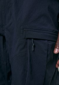 Marineblaue Cargo-Hosen mit glatter Textur, ausgestattet mit einer Reißverschlusstasche, verstellbarem Kordelzug und kontrastierenden Nähten.
