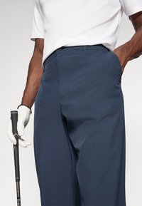 Mornarske golf hlače s elegantnim dizajnom, s ravnim prednjim dijelom, bočnim džepovima i glatkom teksturom. Uparene s bijelim polo majicom i golf rukavicom.