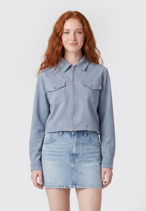 Jonge vrouw met lang rood haar, een lichtblauw jack met rits en een lichtgewassen denim rok, staand tegen een effen achtergrond.