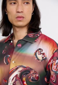 Chemise à motifs présentant un design de bulles brillantes dans des teintes de rouge, vert et marron. Fabriquée en tissu lisse avec un col et des boutons.