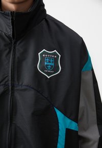 Black jacket with teal and gray accents. Features a shield logo with "BOTTER" and "ANTWERP" text. Smooth, lightweight material. Zippered collar.