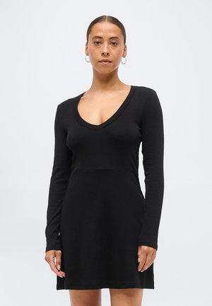 Kvinde med trukket hår tilbage iført sølv hoop-ører og en langærmet sort V-hals kjole mod en ensartet lys baggrund.