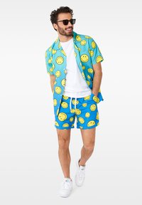Chemise à manches courtes bleue et short assorti avec un motif de smiley. Comprend un t-shirt blanc en dessous et des baskets blanches.