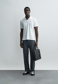 Homme portant un polo blanc et un pantalon gris, tenant une mallette noire, debout devant un fond uni gris clair.