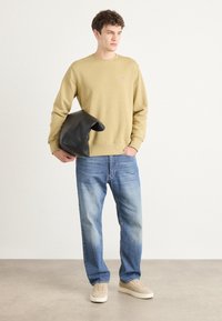 Khaki sweatshirt, losse pasvorm, gecombineerd met lichtblauwe jeans. Houdt een zwarte leren tas met een opvouwbaar ontwerp vast. Beige sneakers.