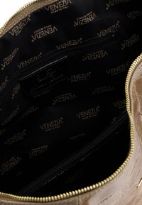Indvendig på en beige kunstlædertaske med sort stoffoder; guld "VENEZIA MADE IN ITALY" mønster; en lynlåslomme og guld lynlåshardware.