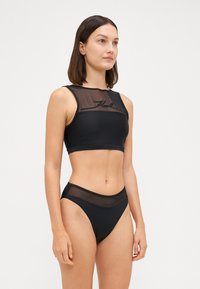 Melnā divdaļīgā peldkostīma komplekts, kas sastāv no bezpiedurkņu topa ar caurspīdīgiem acu akcentiem, apvienots ar augstā jostasvietā esošām bikini apakšdaļām.