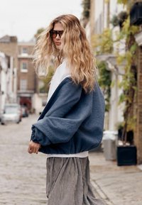 Jachetă supradimensionată din fleece albastru texturat, cu manșete elastice, purtată peste o cămașă albă, asortată cu pantaloni ușori cu dungi.