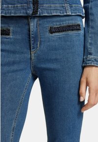 Jeans in denim con un design aderente in blu medio. Dettagli notevoli includono accenti neri testurizzati sulle tasche e cuciture a contrasto.