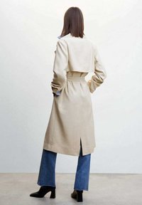 Beiger Trenchcoat mit tailliertem Schnitt, offenem Rücken und seitlichen Schlitzen; kombiniert mit ausgestellten blauen Jeans und schwarzen Stiefeletten.