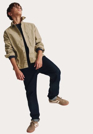 Camicia beige abbottonata, t-shirt nera sotto, jeans scuri e scarpe sportive marroni. Outfit casual, vestibilità comoda e maniche arrotolate.