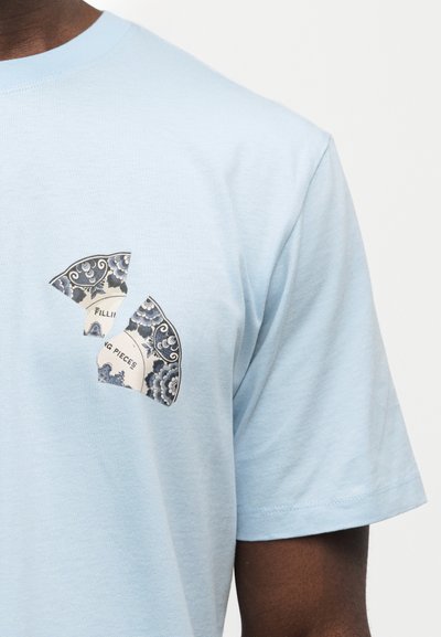 Ανοιχτό μπλε μπλουζάκι με σχέδιο σπασμένου κεραμικού πιάτου και μερικό κείμενο "Filling Pieces" στο στήθος.