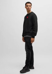 Svart sweatshirt med rund halsringning, prydd med en liten röd logotypapplikation på bröstet. Bärs tillsammans med svarta jeans och svarta sneakers.