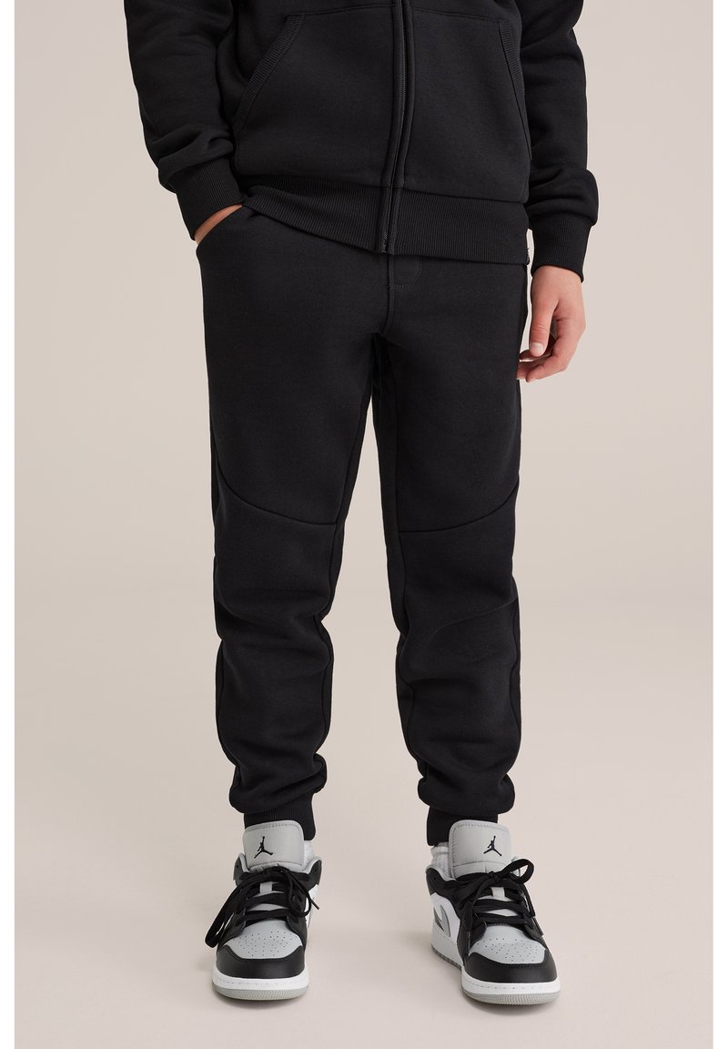 Osoba ubrana w czarne spodnie dresowe i czarno-białe trampki, stojąca na neutralnym, jednolitym tle.