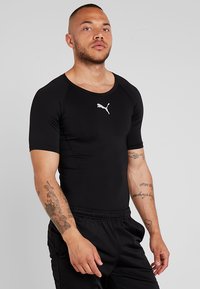 Svart treningstrøye med korte ermer, laget av tettsittende elastisk stoff. Har en hvit Puma-logo på brystet og rund hals.