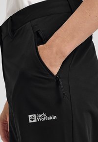 Zwarte outdoorbroek met een gladde textuur, voorzien van een zijzak en het "Jack Wolfskin" logo in het wit. Elastische tailleband met trekkoord.