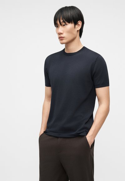 Jeune homme aux cheveux noirs et raides portant un T-shirt à manches courtes bleu marine uni et un pantalon marron foncé, les mains dans les poches.