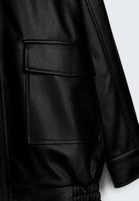 Zwarte leren jas close-up met een grote zak, gestructureerd oppervlak en gathered bottom hem. Gladde afwerking met een elegante uitstraling.