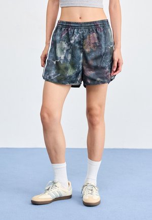 Vrouw draagt camouflageprint sportshorts, witte crew socks en beige Adidas sneakers, staand op een blauwe vloer.
