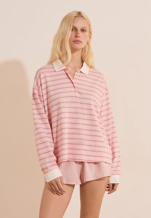 Donna bionda che indossa una camicia rosa a righe a maniche lunghe, larga, con colletto e polsini bianchi, abbinata a pantaloncini rosa chiaro su uno sfondo semplice.