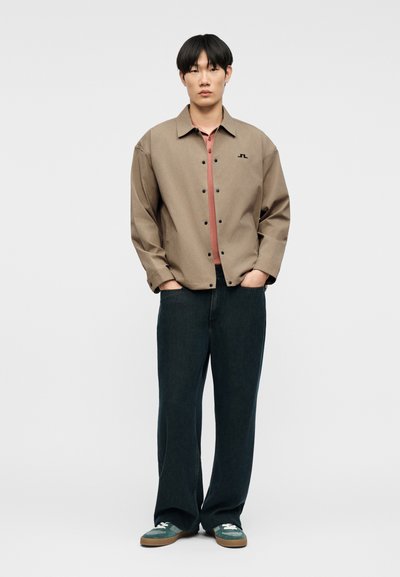 Νεαρός άνδρας στέκεται με τα χέρια στις τσέπες, φορώντας μια μπεζ ζακέτα με κουμπιά πάνω από ένα σκουροκόκκινο μπλουζάκι, μαύρο φαρδύ παντελόνι και πράσινα αθλητικά παπούτσια.