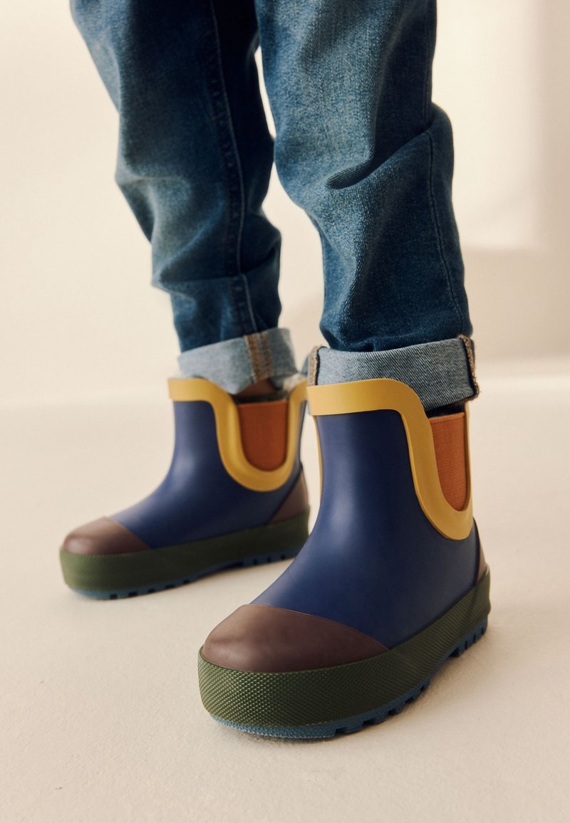 Enfant portant des bottes de pluie colorées avec des détails bleus, bruns, jaunes et verts, associées à un jean retroussé sur un sol clair.