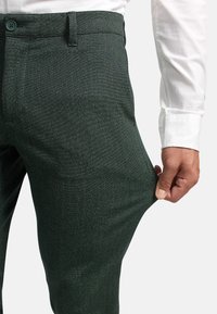 Grüne Hosen im Fischgrätmuster aus strukturiertem Stoff, mit einem Knopfverschluss und einem Handgriff, der den Bund einstellbar zieht.