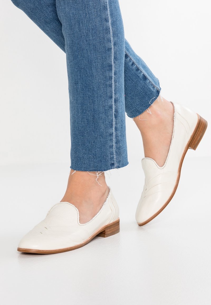 Ballerines en cuir blanc avec un petit talon en bois, présentant des ouvertures courbes et des détails de couture discrets. Portées avec un jean en denim effiloché.